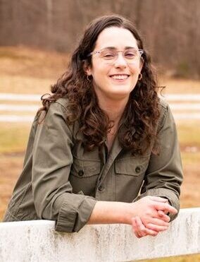 Abigail Murray Person with long curly hair and glasses, wearing a green jacket, smiling and leaning on a wooden fence in an outdoor setting, looking like one of the trusted providers of warmth and cheer.