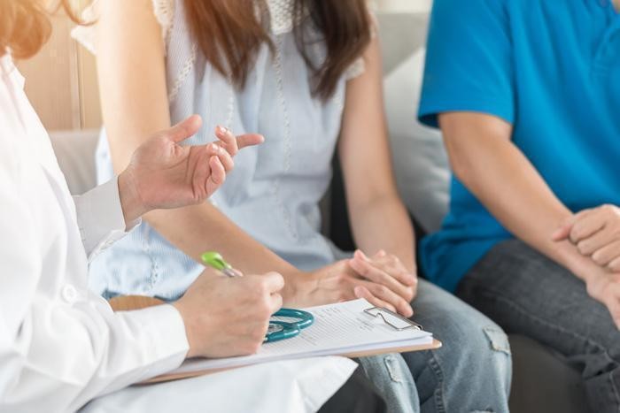 A doctor holds a pen and clipboard during a consultation with two seated patients, one in a light blue sleeveless top and the other in a blue shirt.