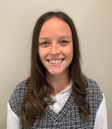 A woman with long, dark hair is smiling at the camera. She is wearing a white shirt with a black and white checkered vest, exuding confidence like one of those trusted providers. The background is plain and light-colored.