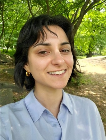 Maya Rubin A person with short dark hair and wearing a light blue shirt smiles in an outdoor setting, surrounded by trees and greenery. They exude warmth, much like trusted providers of care in a community environment.
