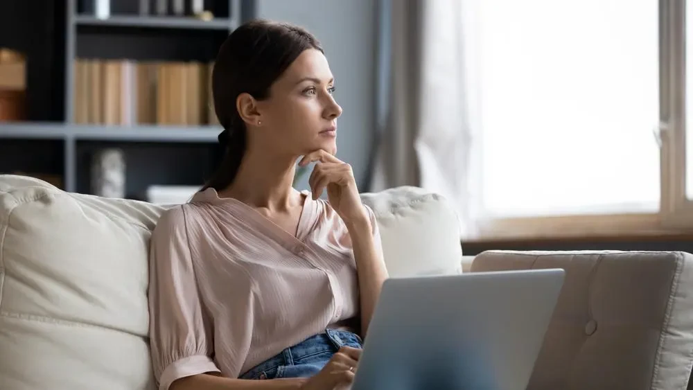 A woman sits on a couch, resting her chin on her hand, and looks thoughtfully into the distance with a laptop on her lap. Shelves with books and decorations are visible in the background.