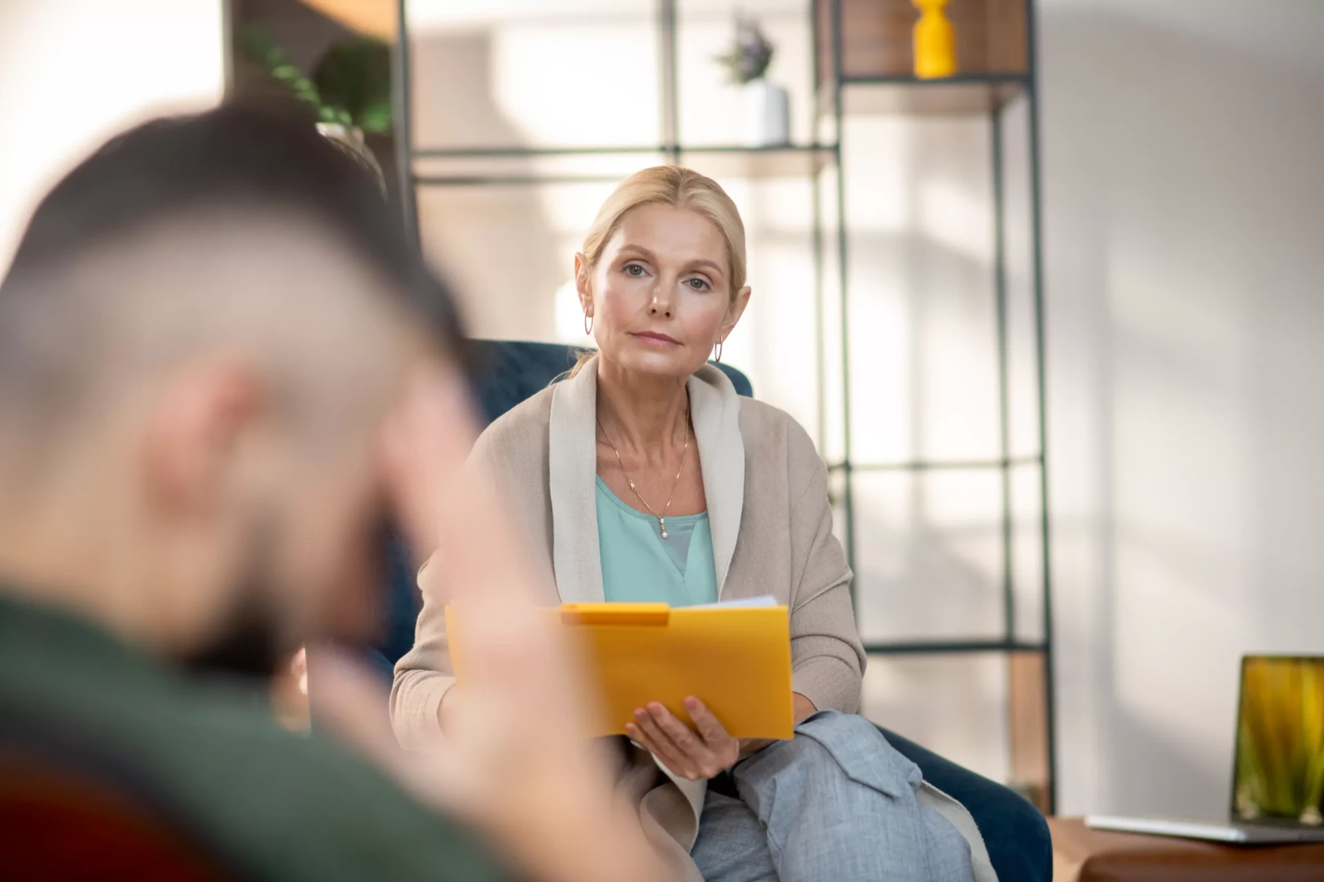A woman holding a yellow folder is seated, attentively listening to a man in the foreground who appears to be talking.