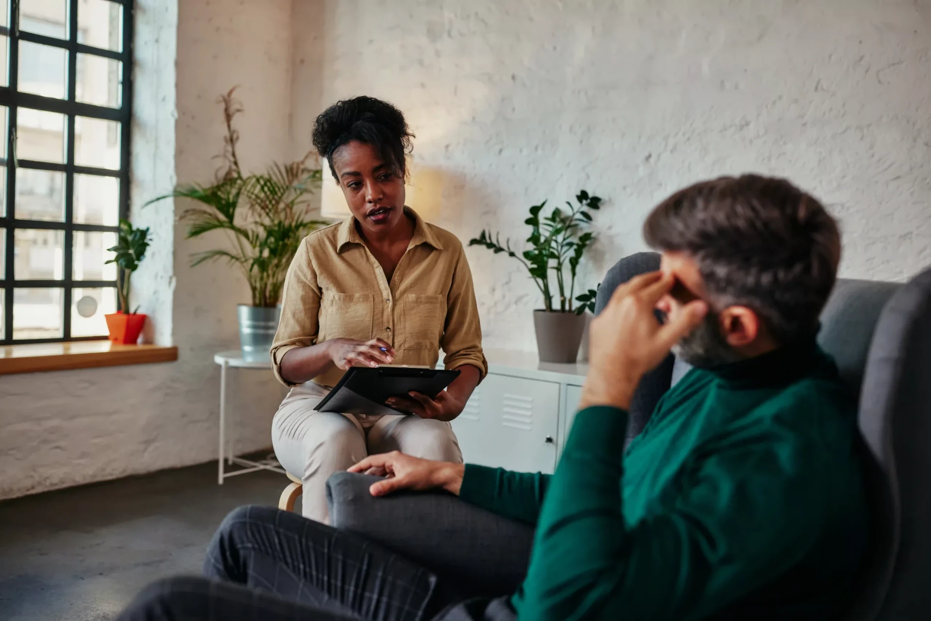 A woman sits with a tablet, talking to a man reclining in a chair in an office setting with potted plants and a window in the background.