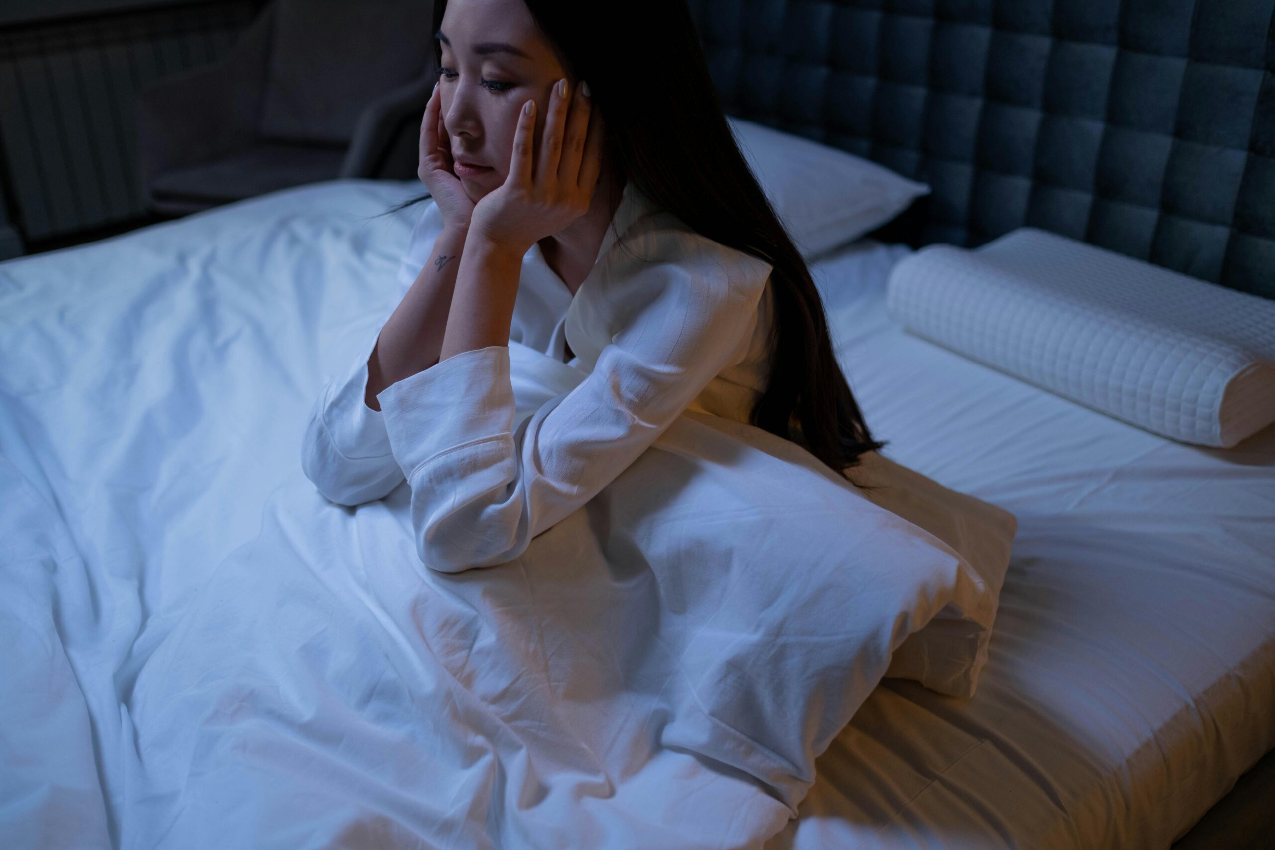 A woman in white pajamas sits on a bed at night, resting her chin on her hands.