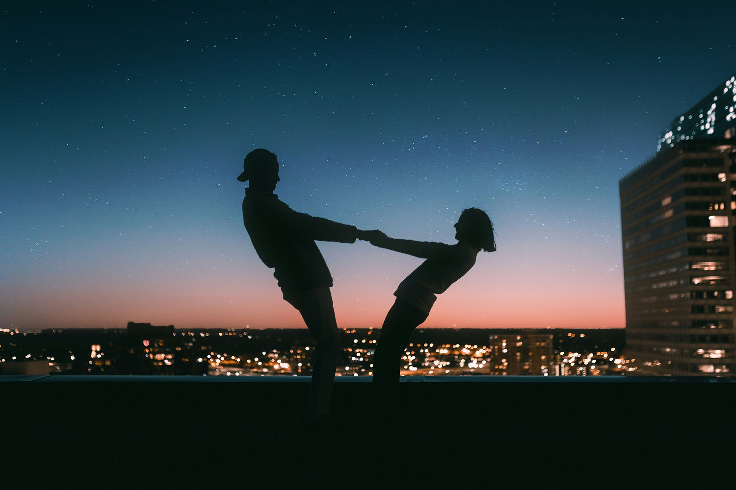 Silhouetted couple holding hands, leaning back against a cityscape as stars twinkle above at dusk, embracing the gentle journey of dating someone with social anxiety.