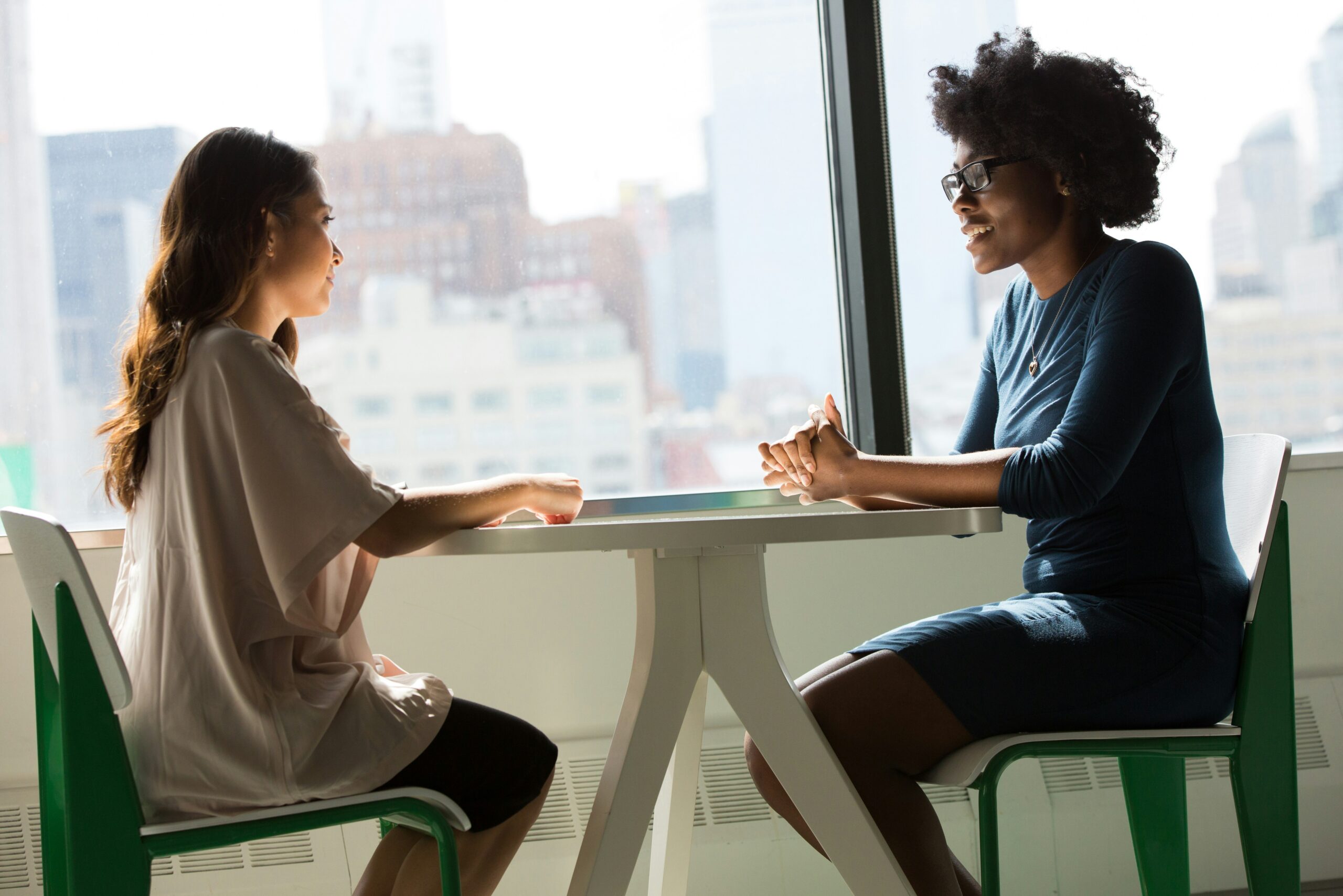 Two women are sitting at a table near a large window, engaged in conversation. The room is brightly lit by natural light.
