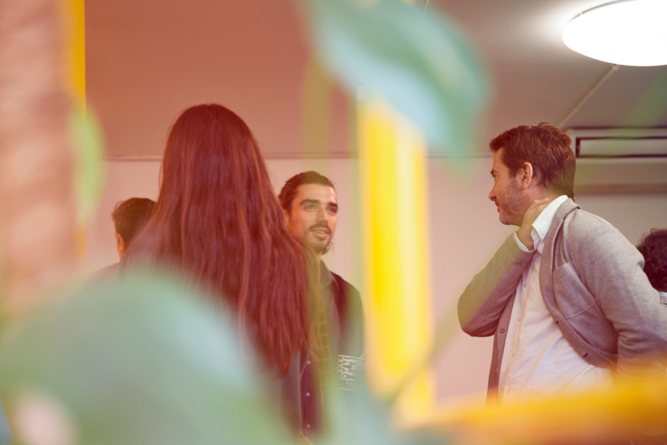 Three people standing and talking in an indoor setting, with a blurred plant in the foreground.