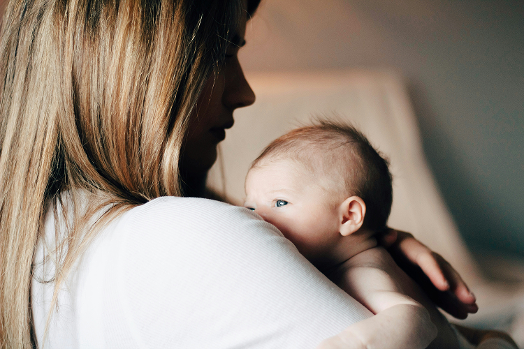 A woman holding a baby against her shoulder. The baby is looking to the side, and both are in a softly lit room.