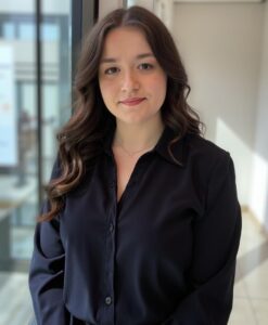 Elizabeth Malakhova A person with long dark hair, reminiscent of Alexa Stern, wearing a black shirt, stands by a window indoors, bathed in natural light.