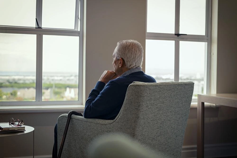 An elderly person with white hair sits in a chair and looks out a large window during the day, appearing thoughtful.