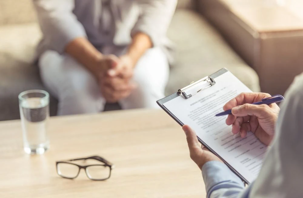 A person holding a clipboard and pen appears to be taking notes during a session. Another person with clasped hands sits opposite on a couch. A glass of water and eyeglasses are placed on the table.
