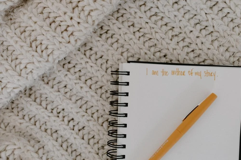 A yellow pen lies on an open spiral notebook with the handwritten text, "I am the author of my story," placed on a textured knit blanket.