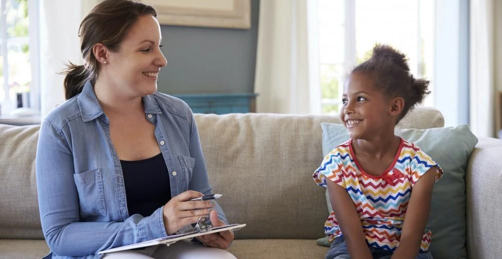An adult and a child sit on a couch indoors, both smiling at each other. The adult holds a clipboard and pen.