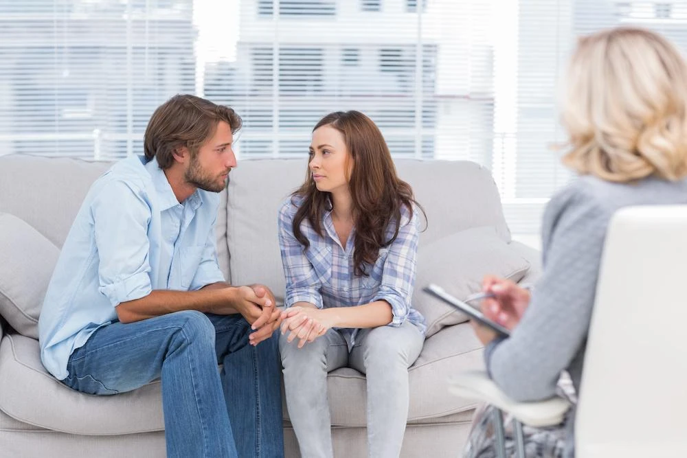 A couple sits closely on a sofa, looking at each other intently while a therapist, sitting in a chair with a clipboard, observes them. The setting appears to be a counseling office.