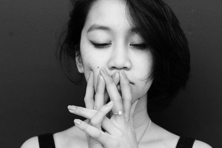 Black and white photo of a person with eyes closed, hands near their face, fingers intertwined. They have shoulder-length hair and wear a sleeveless top.