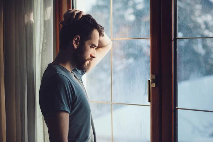 A man with a beard and dark hair stands by a window, looking outside with one hand on his head. He appears thoughtful, with natural light streaming in through the window.