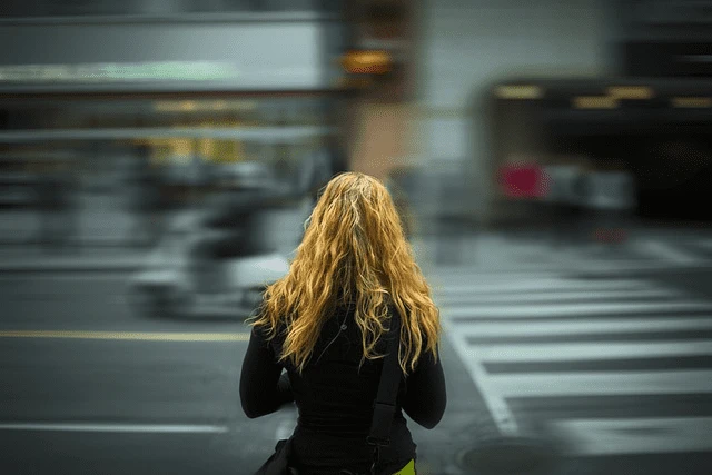 A person with wavy blonde hair stands at a crosswalk in a busy city street, facing away from the camera, embodying an aura of quiet depression amidst the urban hustle.