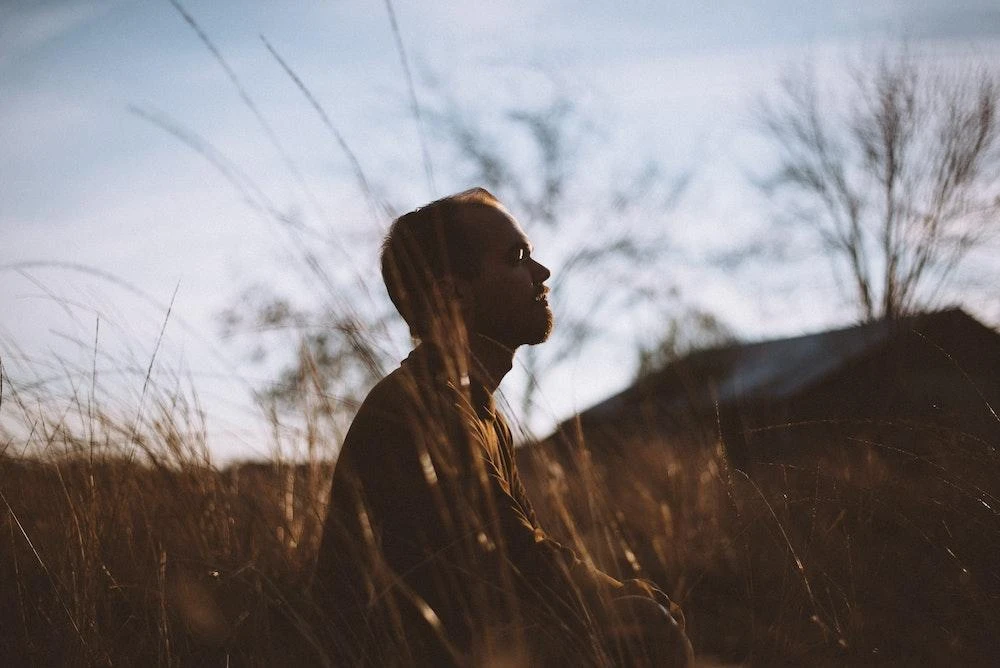 A person sits quietly in a field at sunset, with tall grass around them and a barn or structure in the blurred background.
