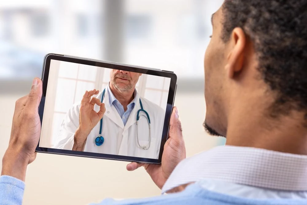 A person is holding a tablet showing a video call with a doctor, who is making an "OK" gesture while wearing a white coat and stethoscope.