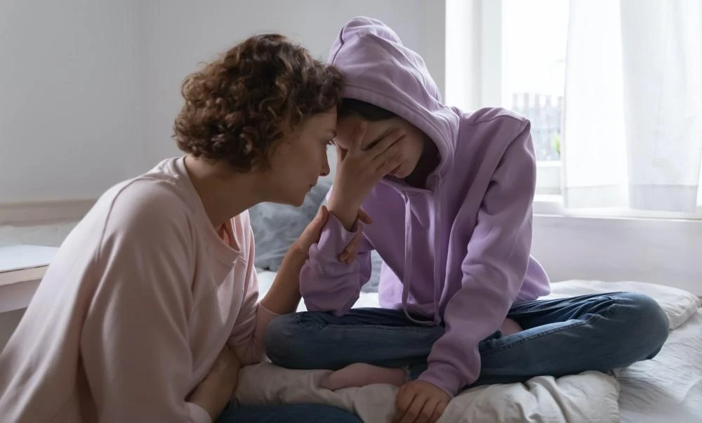 A person consoles another person who is sitting on a bed, wearing a purple hoodie and holding their head in their hand.