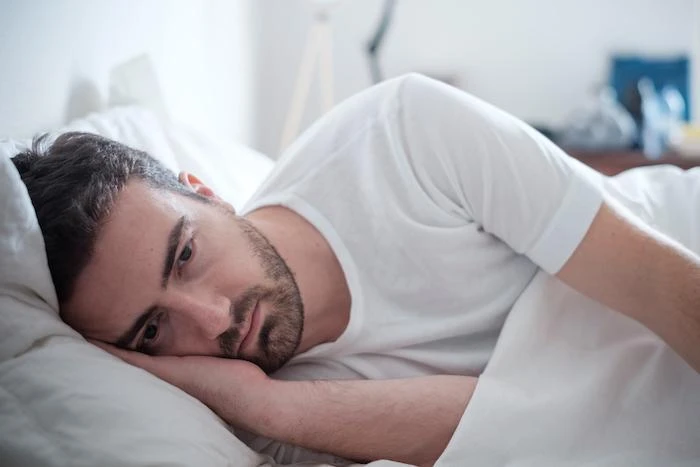 A man lies in bed on his side, resting his head on a pillow and looking ahead with a neutral expression, wearing a white t-shirt.