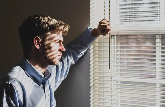 A man in a button-up shirt stands by a window with closed blinds, his arm resting on the frame, as he looks outside with a contemplative expression.