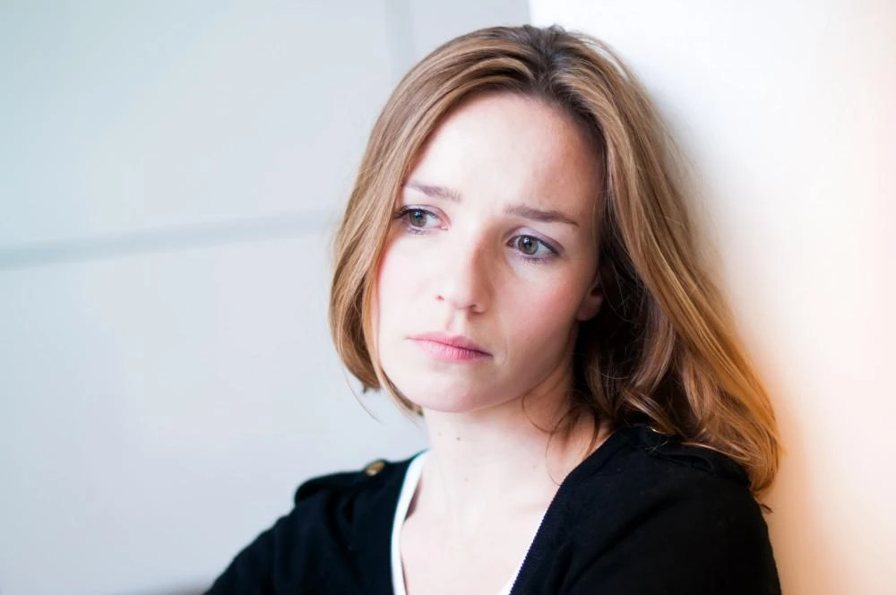 A woman with long brown hair leans against a white wall, looking into the distance with a thoughtful expression.