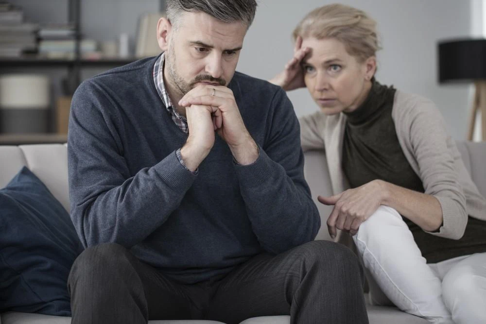 A man and a woman sit on a couch, both appear worried and deep in thought. The man has his hands clasped in front of him, while the woman rests her hand on her forehead.