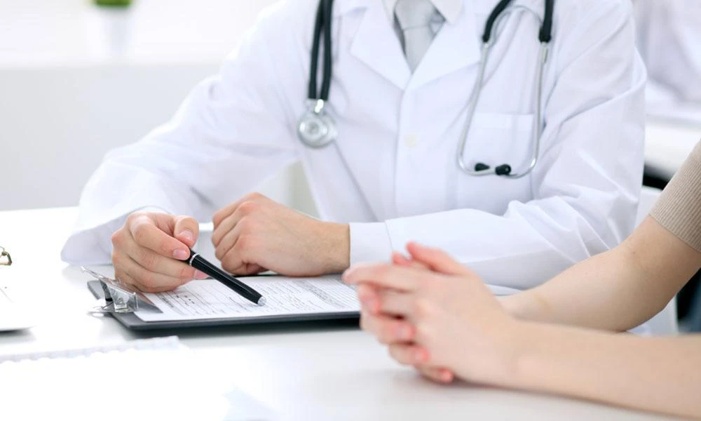 A doctor wearing a white coat and stethoscope points at a document with a pen while another person sits across with hands clasped.
