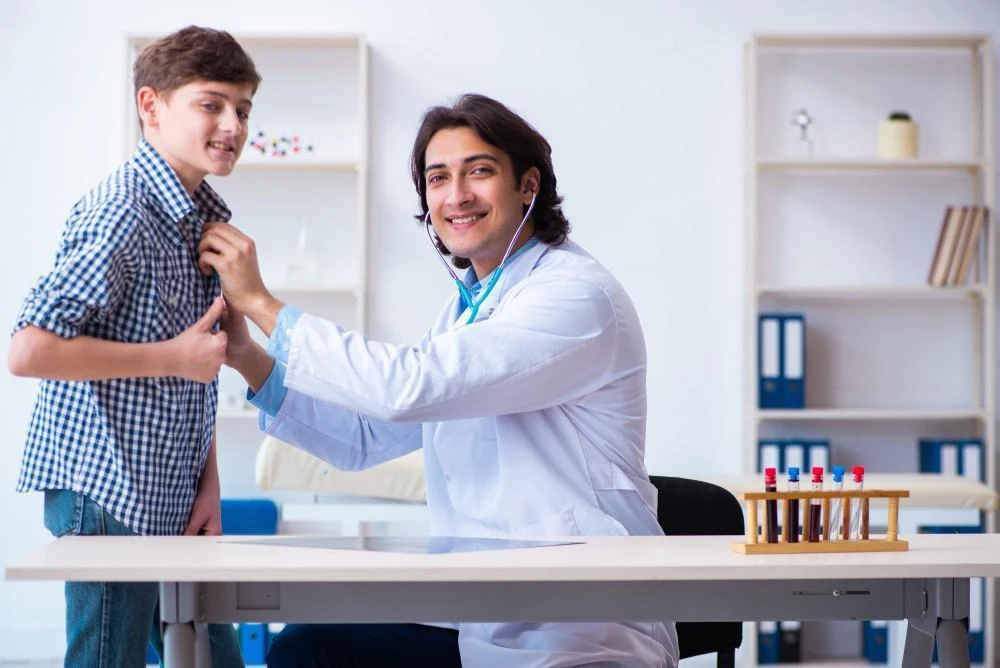 A doctor uses a stethoscope to examine a boy in a checkered shirt, who gives a thumbs-up. They are in a medical office setting.