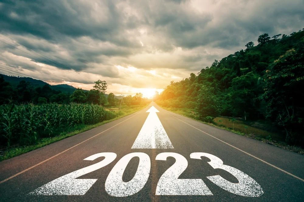 A road with a large "2023" and a white arrow painted on it stretches into the distance towards the horizon under a cloudy sky, flanked by greenery.