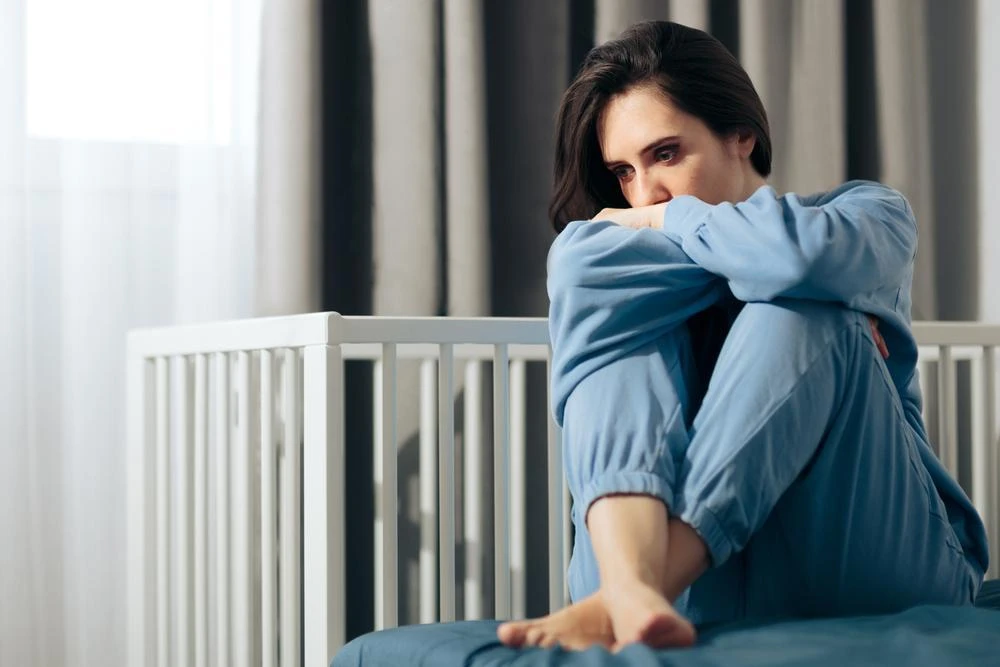 A woman wearing blue sits on a bed hugging her knees, looking pensive, with a crib in the background.