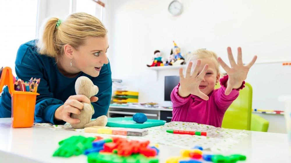 A woman with a blond ponytail observes a young girl who is showing her open palms. They are seated at a table with various colorful toys and art supplies in a bright, airy room.
