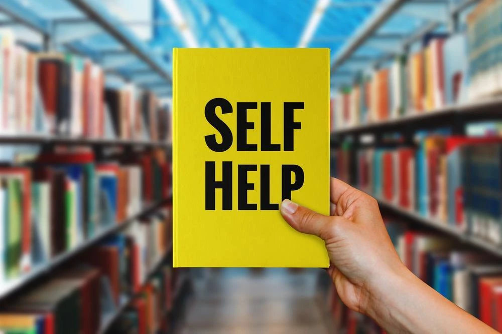 A person holds a yellow book titled "SELF HELP" in front of bookshelves in a library.