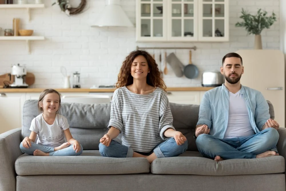 A woman, man, and child sit on a sofa in a meditative pose in a modern kitchen. The woman and man have their eyes closed, while the child smiles with eyes open.