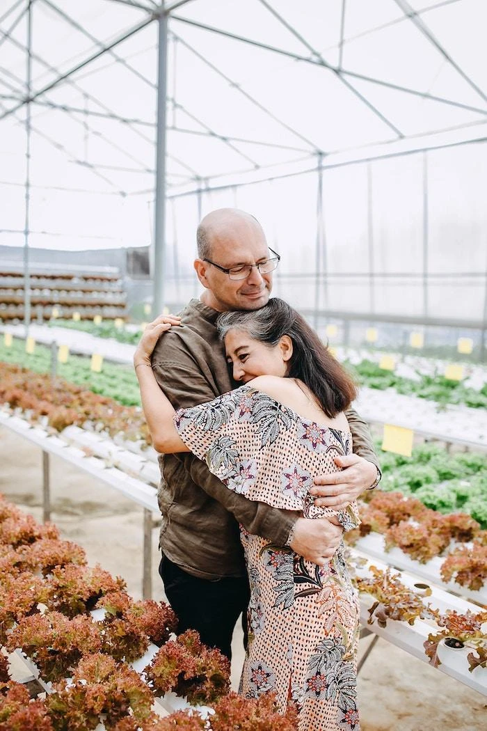 A man and a woman embrace each other in a greenhouse filled with rows of leafy plants.