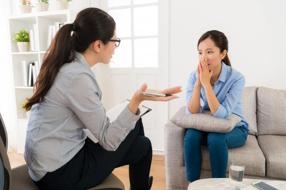 Two women sit facing each other. One woman is holding a phone and gesturing while the other looks thoughtful with her hands clasped near her face. They appear to be in a serious conversation.