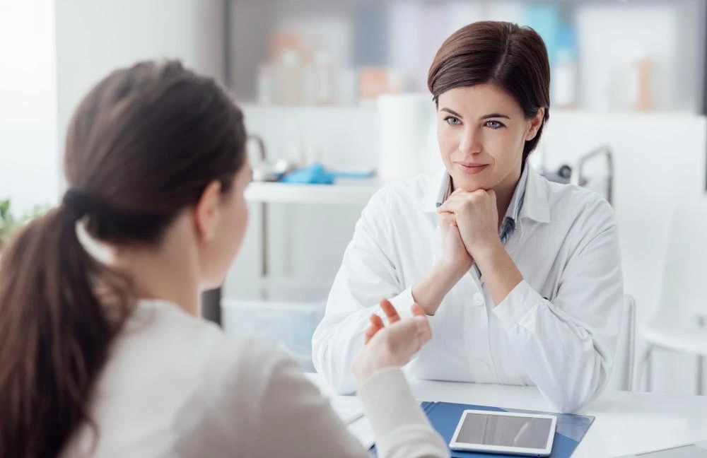 A doctor attentively listens to a patient during a consultation, holding her hands under her chin. The patient is gesturing while they talk. An electronic device is visible on the desk.