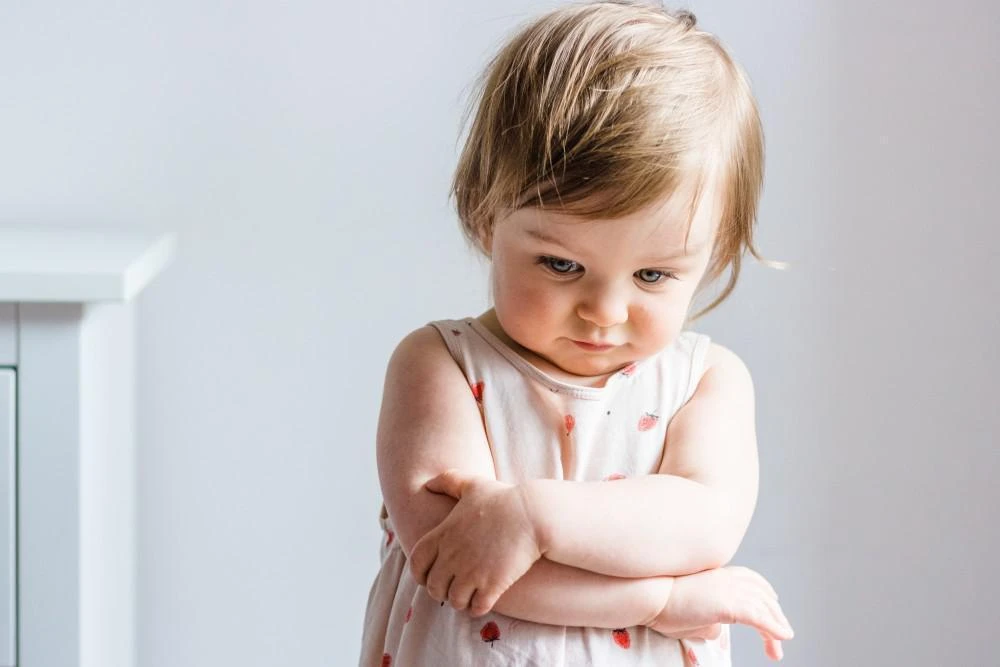 A toddler with light brown hair stands indoors, looking down with arms crossed, wearing a light-colored dress with small red patterns.