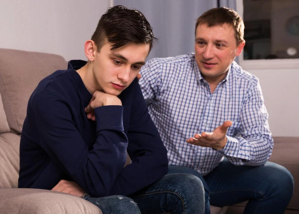 A man in a checked shirt talking to a sad-looking teenage boy who is sitting with his head resting on his hand.