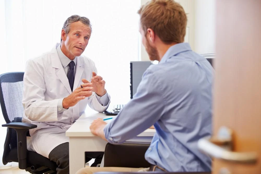 A doctor in a white lab coat is sitting at a desk and talking to a man in a blue shirt, who is sitting across from him. The doctor is gesturing with his hands during the conversation.