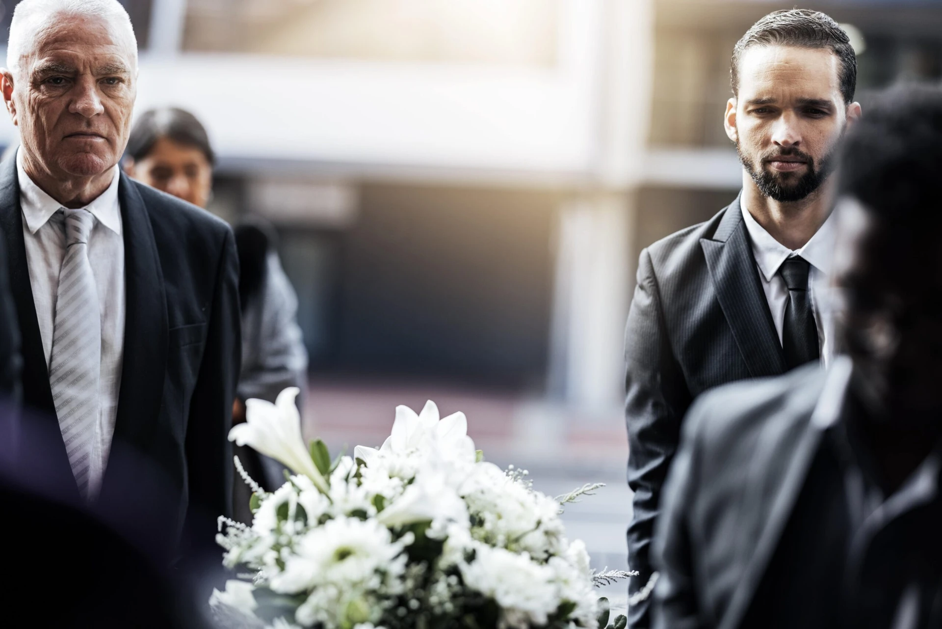 A group of people in formal attire are at an outdoor daytime event. One person is holding a wreath of white flowers.