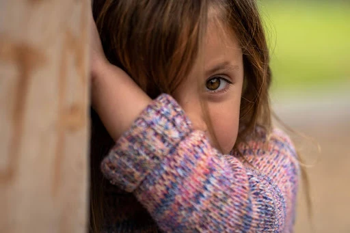A young child with long hair and brown eyes looks cautiously from behind her hand, with one eye partially covered. She is wearing a multi-colored knit sweater.