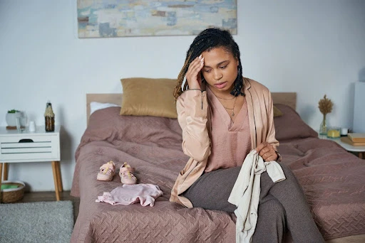 A person sits on a bed, holding clothes and touching their head with a pensive expression. Children's shoes and clothing are nearby.