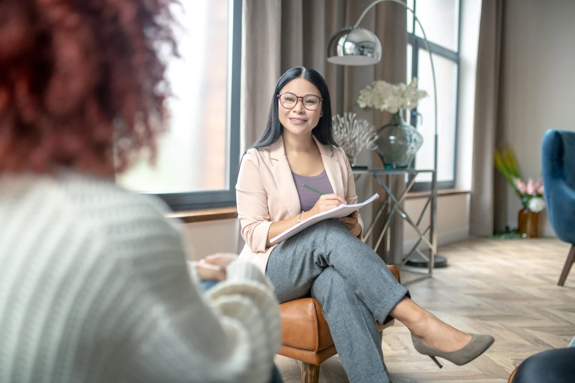 A woman in glasses and a beige blazer sits with a notepad, listening attentively to another person in a cozy room.