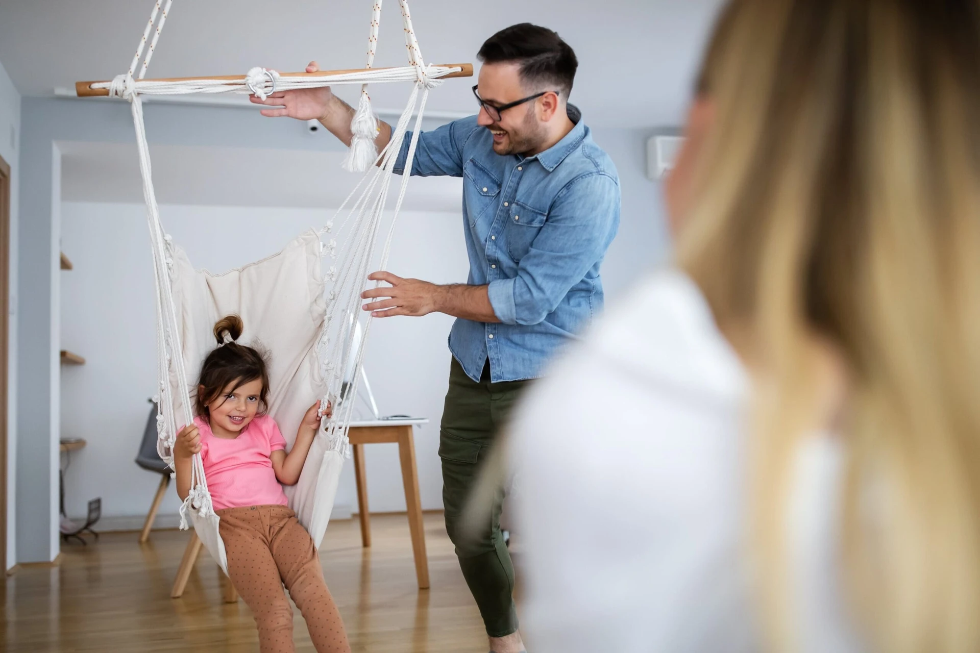 A man playfully pushes a smiling child in a hanging swing indoors, while a woman watches in the foreground.