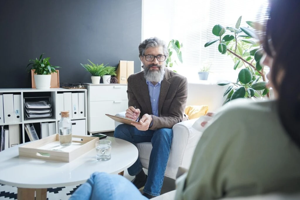 A bearded man in glasses writes on a notepad while sitting on a couch in a modern office with plants. Another person is seated across from him.